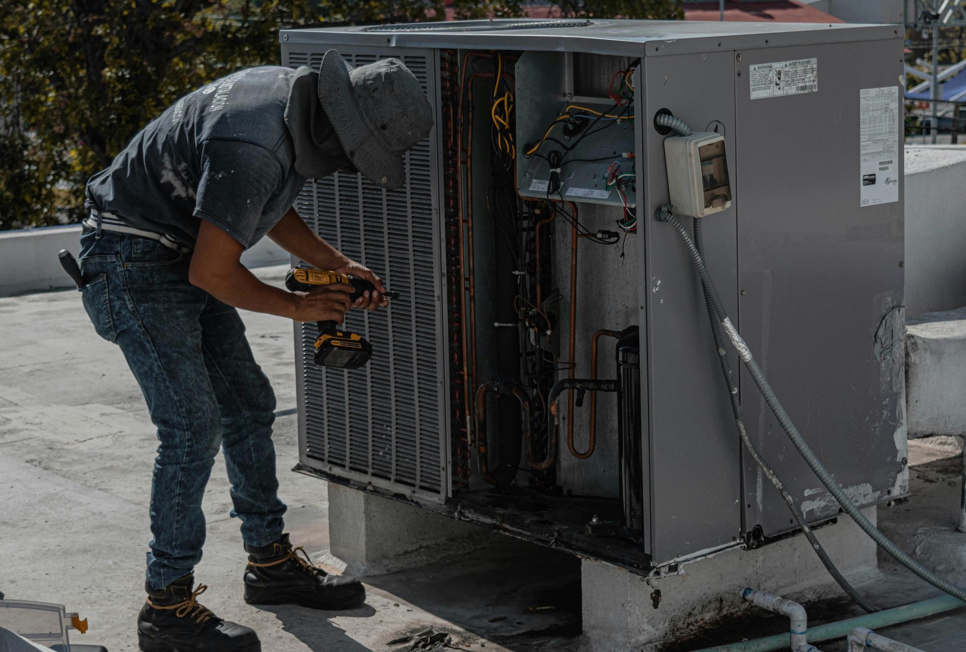 A worker in a bucket hat repairs an outdoor air conditioning unit on a rooftop.